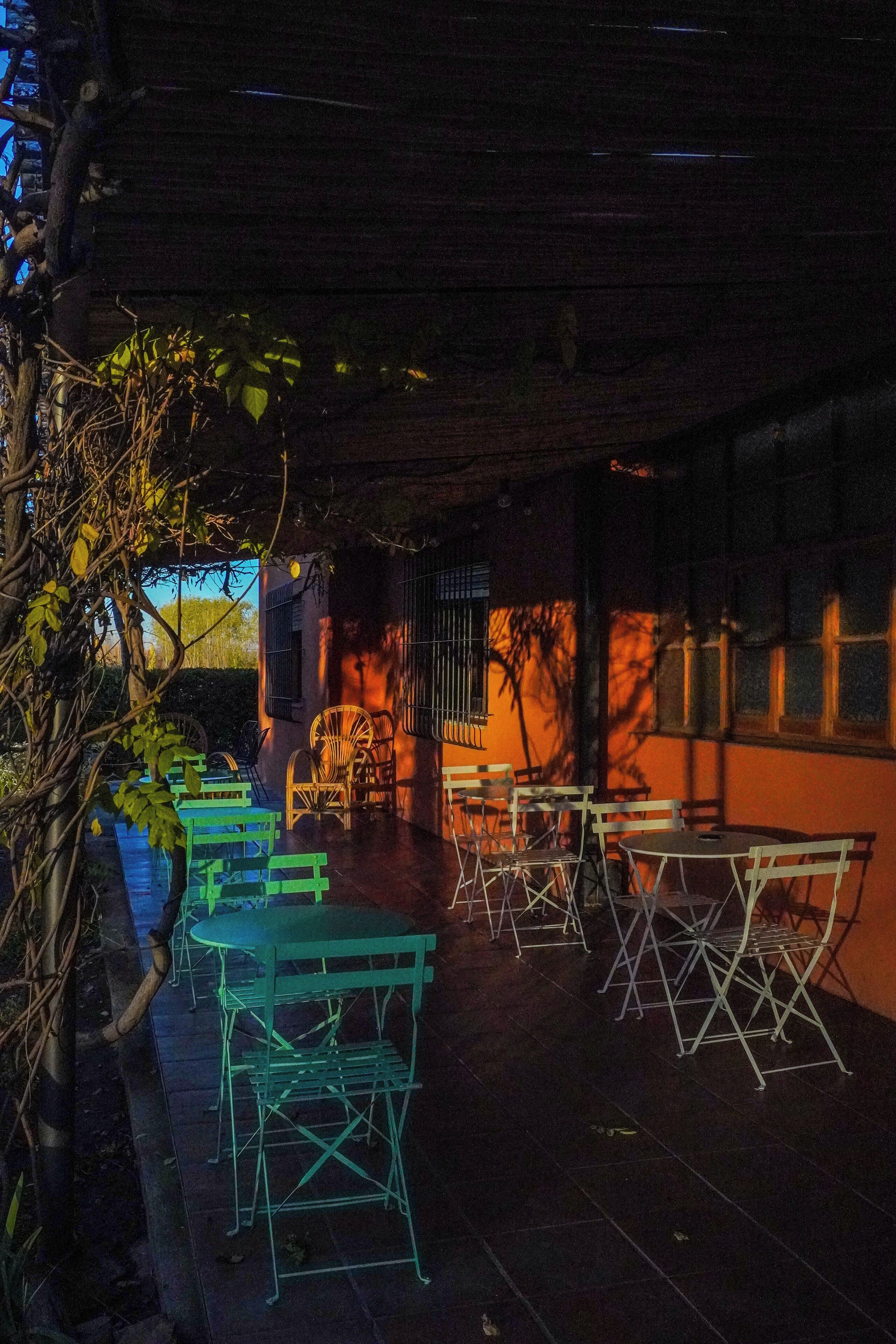 Un patio al aire libre con mesas y sillas de metal, rodeado de árboles y un edificio con ventanas de celosía.