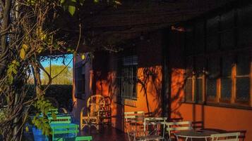 Un patio al aire libre con mesas y sillas de metal, rodeado de árboles y un edificio con ventanas de celosía.