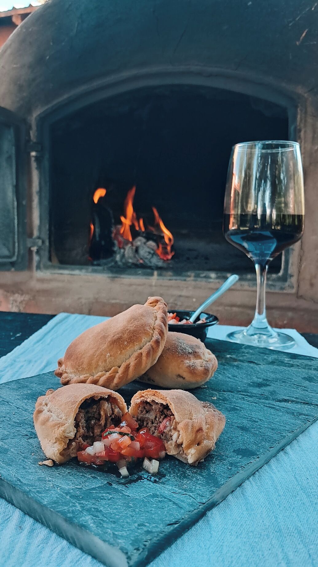 A table with a plate of stuffed pastries and a glass of red wine, set against a brick oven with a fire.