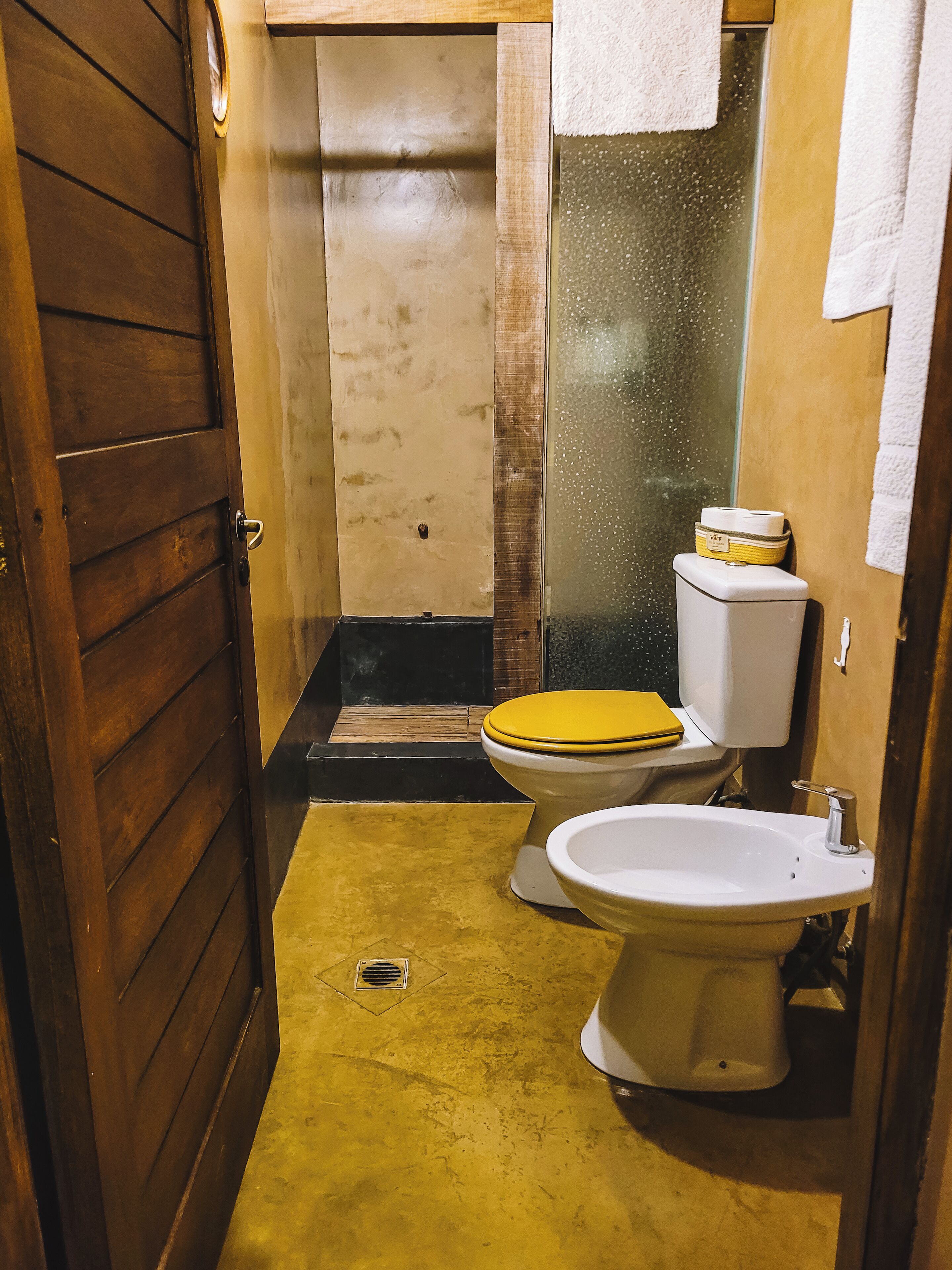 A small, narrow bathroom with a wooden door, a yellow toilet, a white sink, and a towel hanging on the wall.