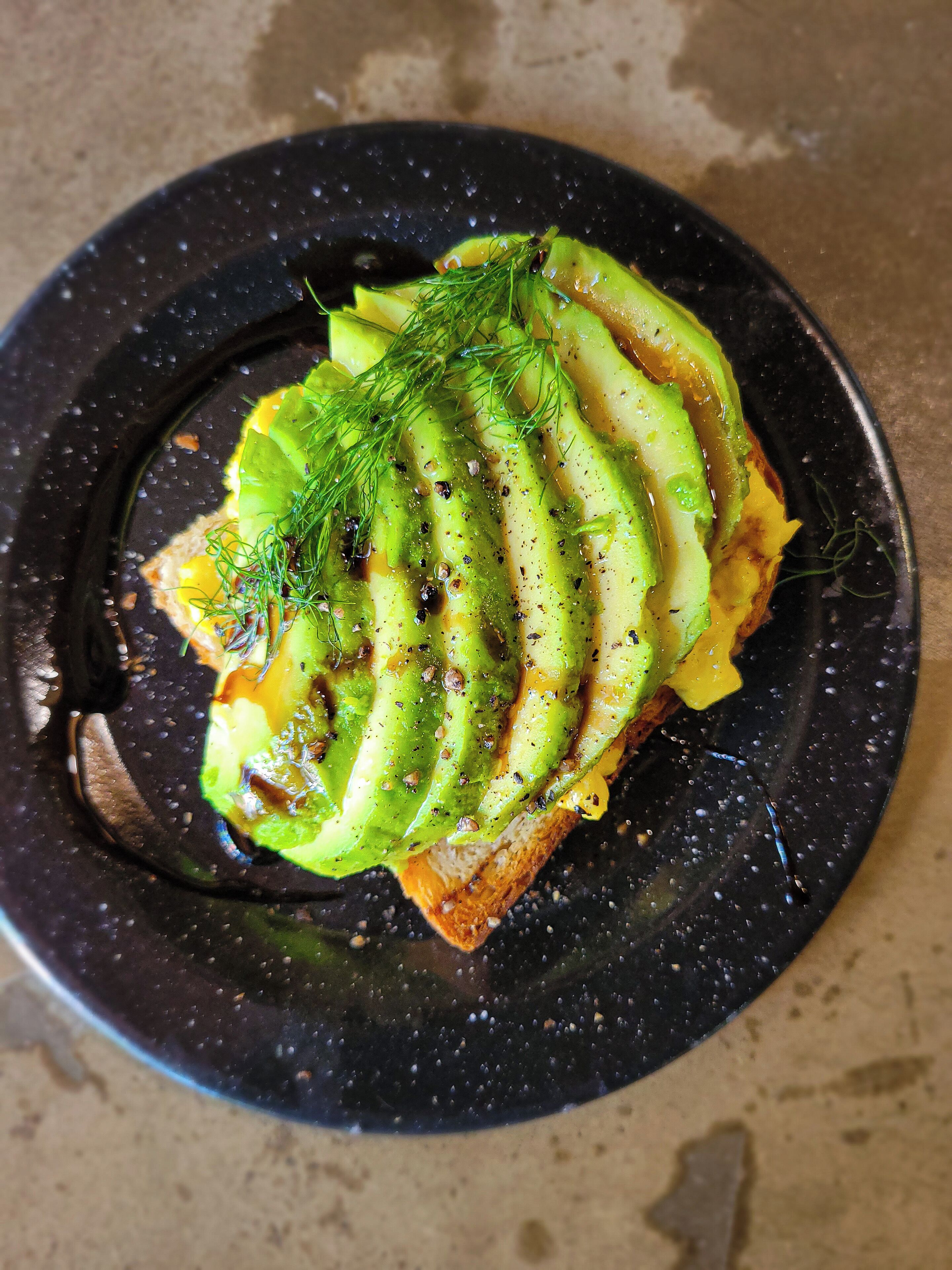 A black plate with a slice of toast topped with avocado slices and dill.