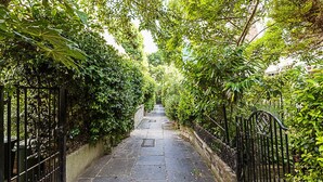 A stone pathway flanked by dense foliage and black metal gates, leading to a building.