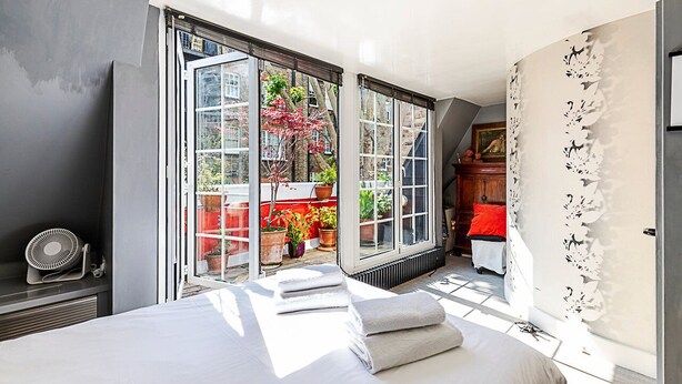 A bedroom with a large bed, a radiator, a fan, and a view of a garden through open French doors.