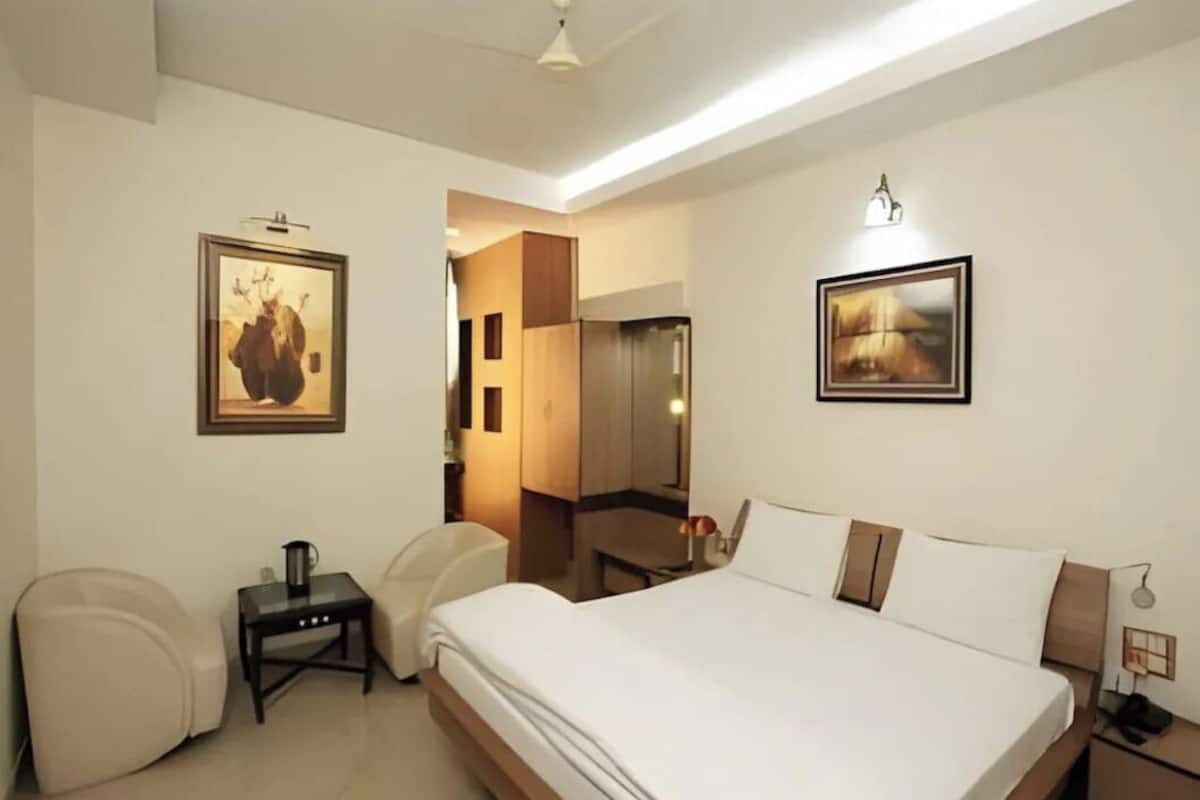 A hotel room with a large bed, a desk, a chair, a small table with a kettle, and two framed pictures on the walls.