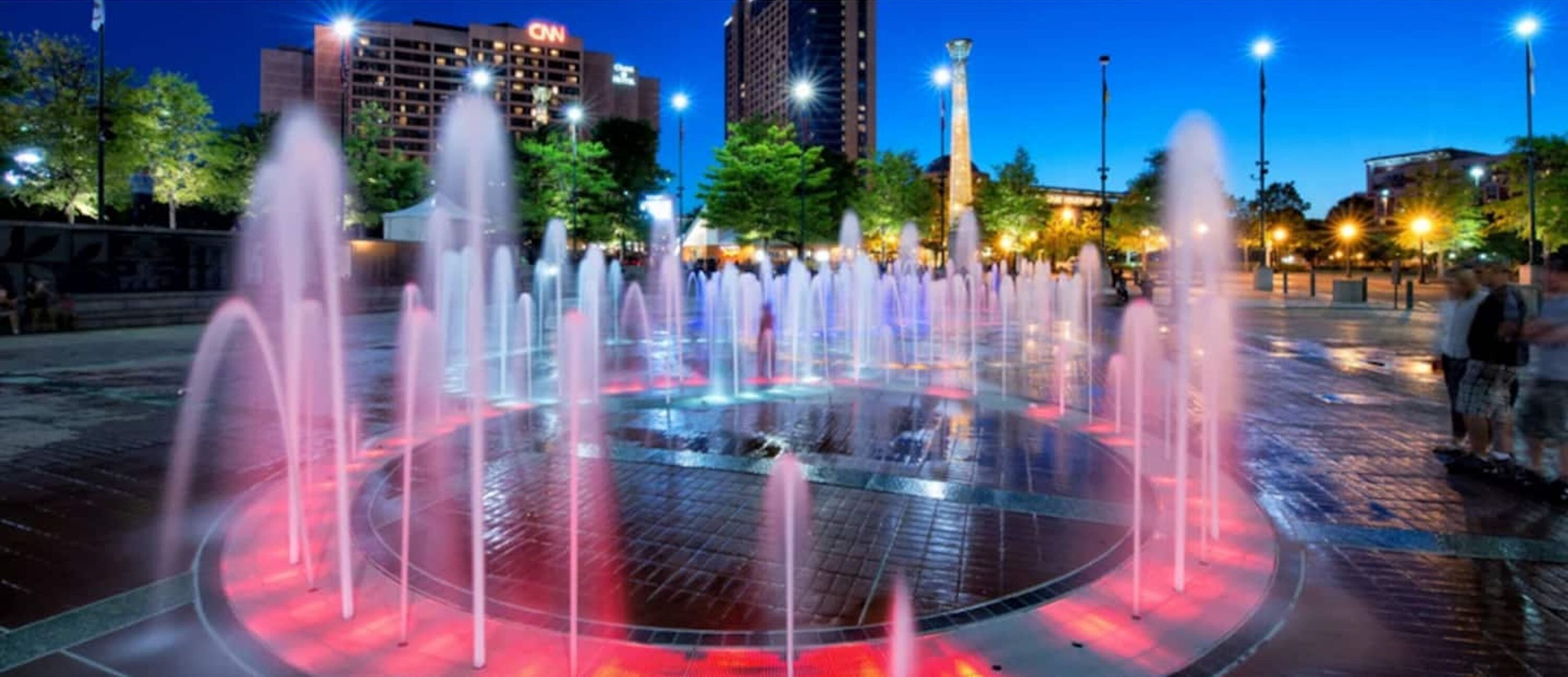 A fountain with multiple water jets in a circular basin, surrounded by a plaza with trees and a building with 'CNN' signage in the background.