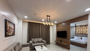 A modern living room with a sofa set, a coffee table, a TV wall unit, and a bedroom with a bed and decorative pillows. - Anandi Bhawan by Pandora, Vrindavan (Mathura)