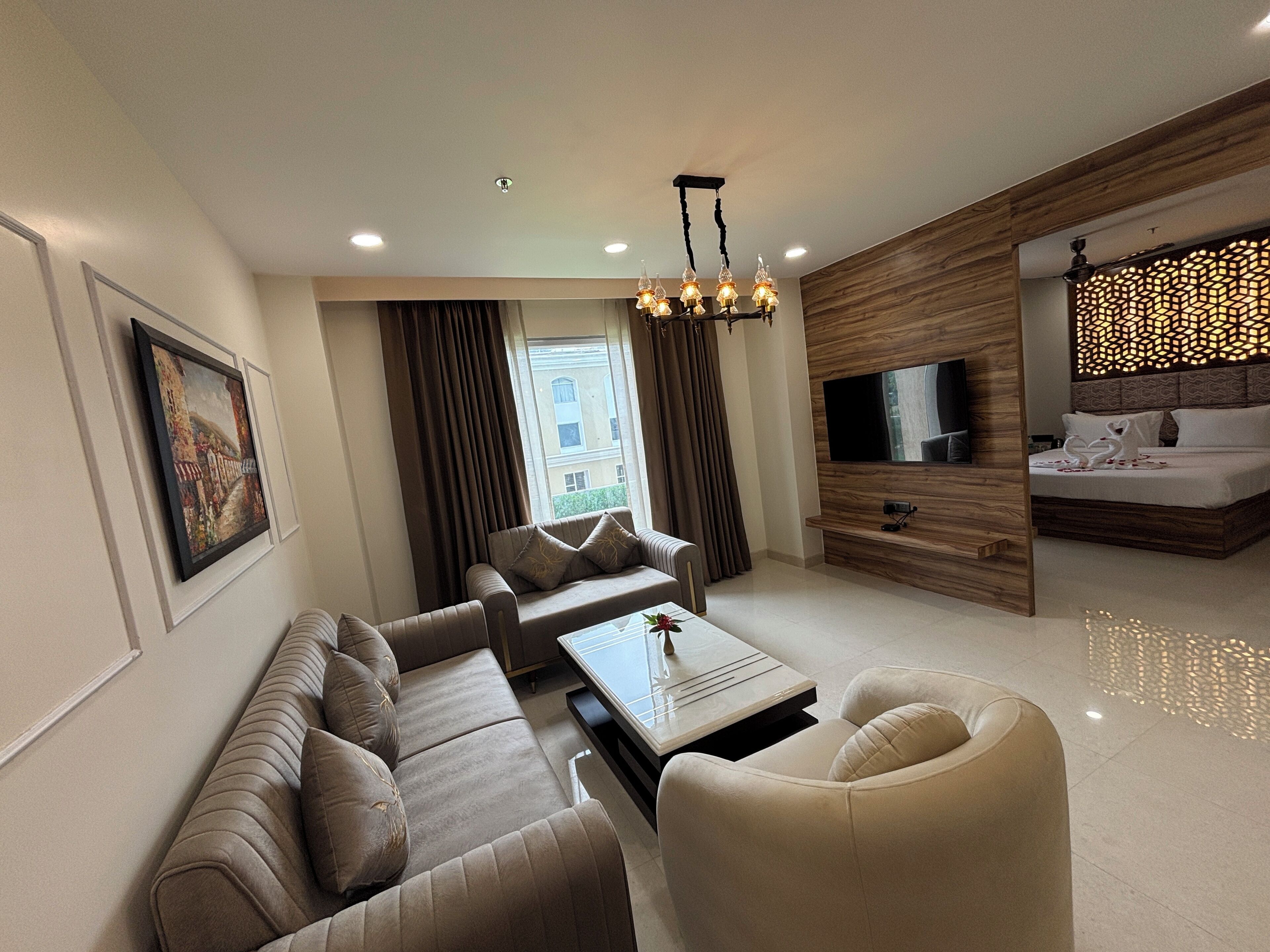 A modern hotel room with a living area, a sofa set, a coffee table, a TV, and a bed with a decorative headboard.