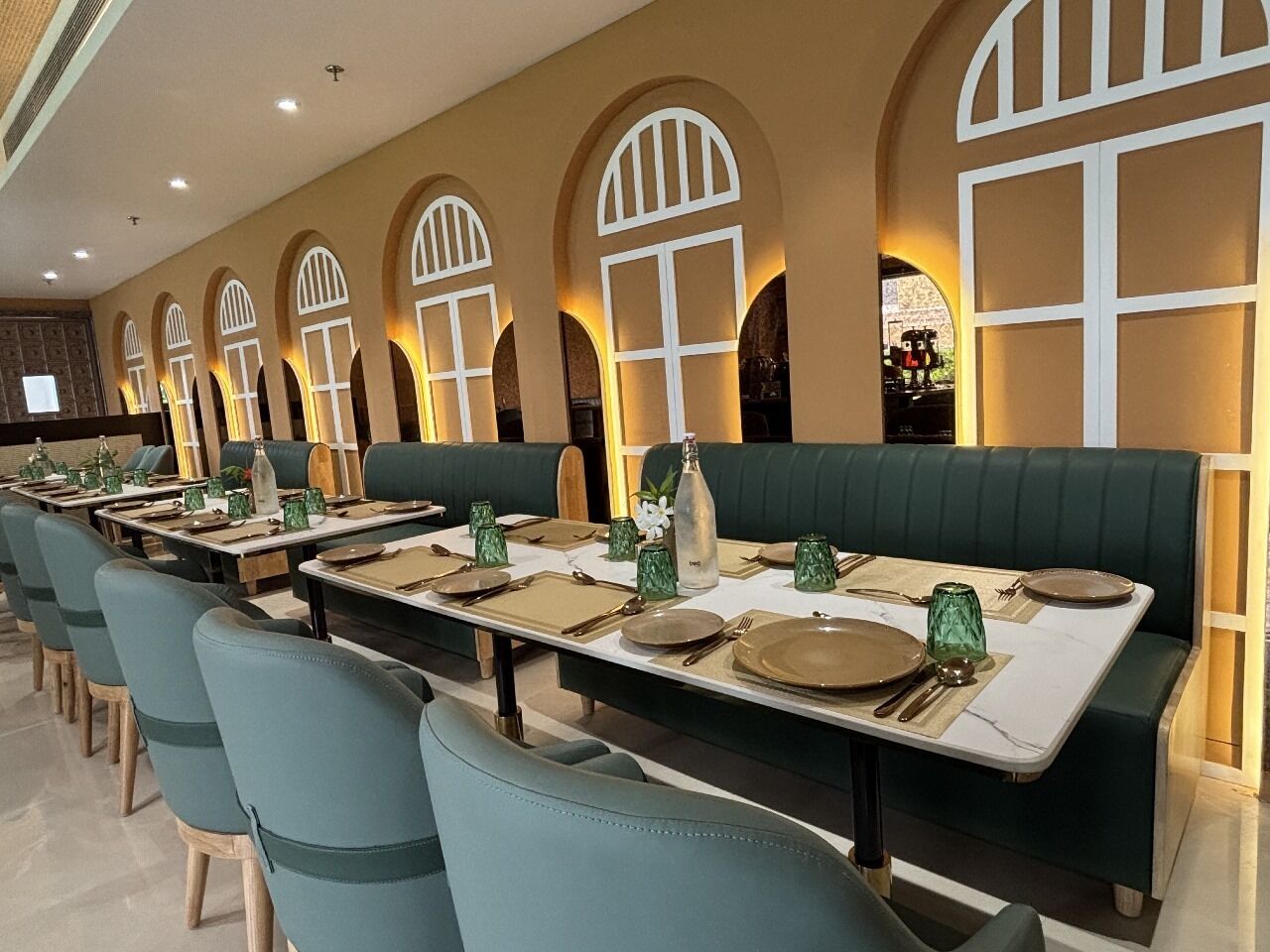 A restaurant with long tables, green cushioned booths, and arched window designs.