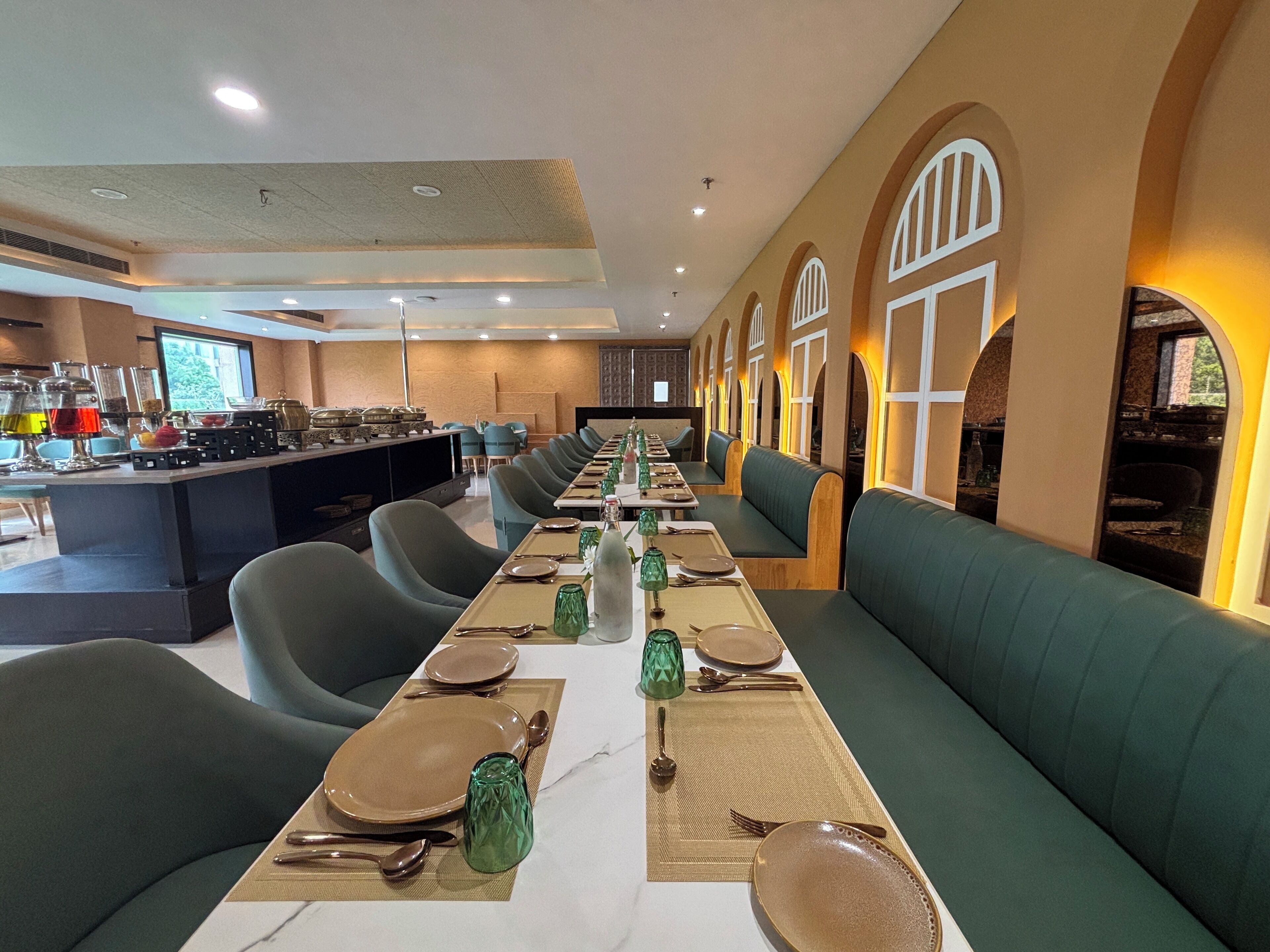Una larga mesa de comedor con asientos acolchados verdes, una zona de bufé en un lateral y ventanas arqueadas a lo largo de la pared.