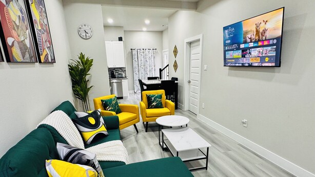 A living room with a green sofa, yellow armchairs, a white coffee table, and a flat-screen TV mounted on the wall.