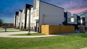 A modern multi-story residential building with a flat roof, large windows, and a wooden fence in front.