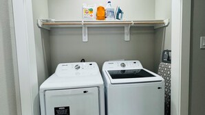 A laundry room with two front-loading washing machines and a shelf above containing cleaning supplies.