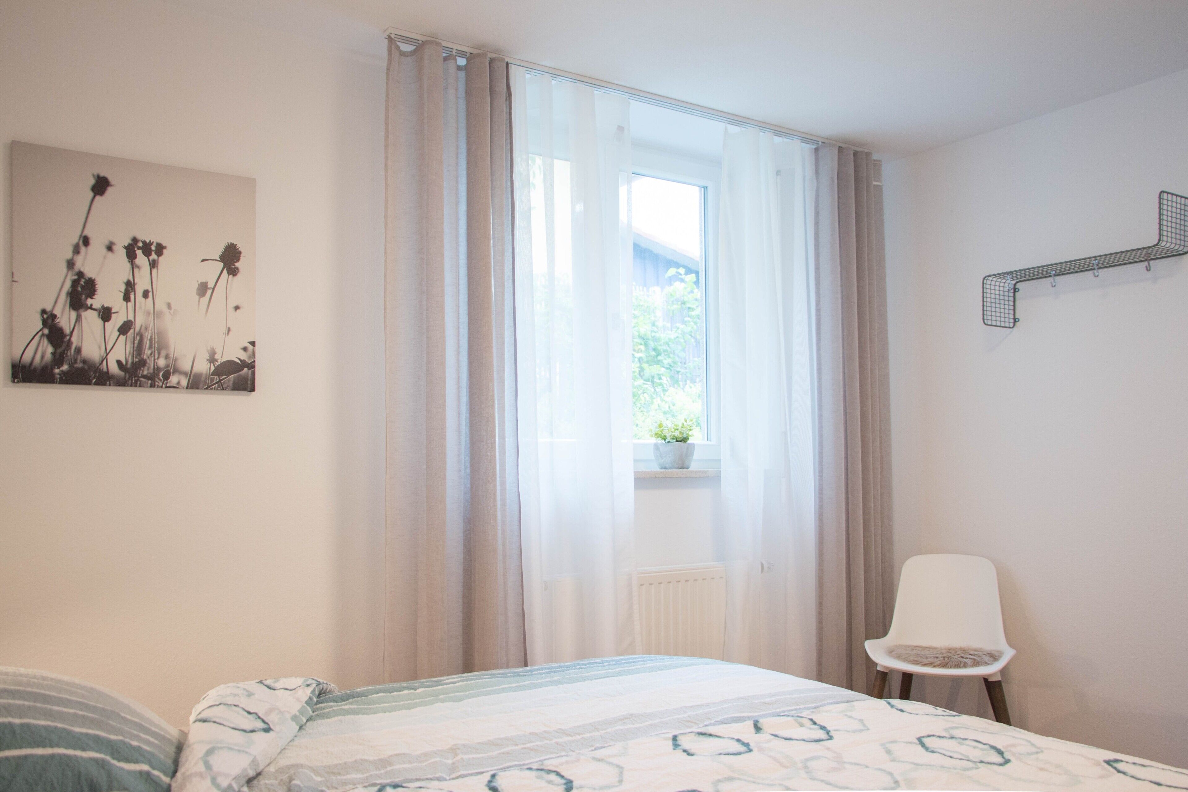 A bedroom with a bed, a chair, a window with curtains, a radiator, and a wall-mounted shelf.