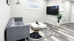 A modern living room with a grey sofa, a white coffee table, a flat-screen TV, and a potted plant. - Beautiful Cozy Spacious Suite (Calgary)