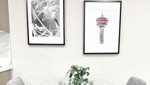 A modern dining area with a glass table, two grey upholstered chairs, and two framed artworks on the wall. - Beautiful Cozy Spacious Suite (Calgary)