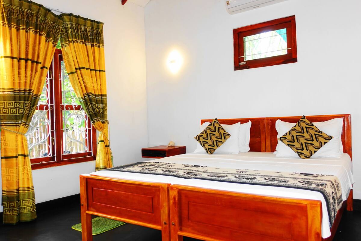 A room with a wooden bed, a window with curtains, a bedside table, and a wall-mounted light.
