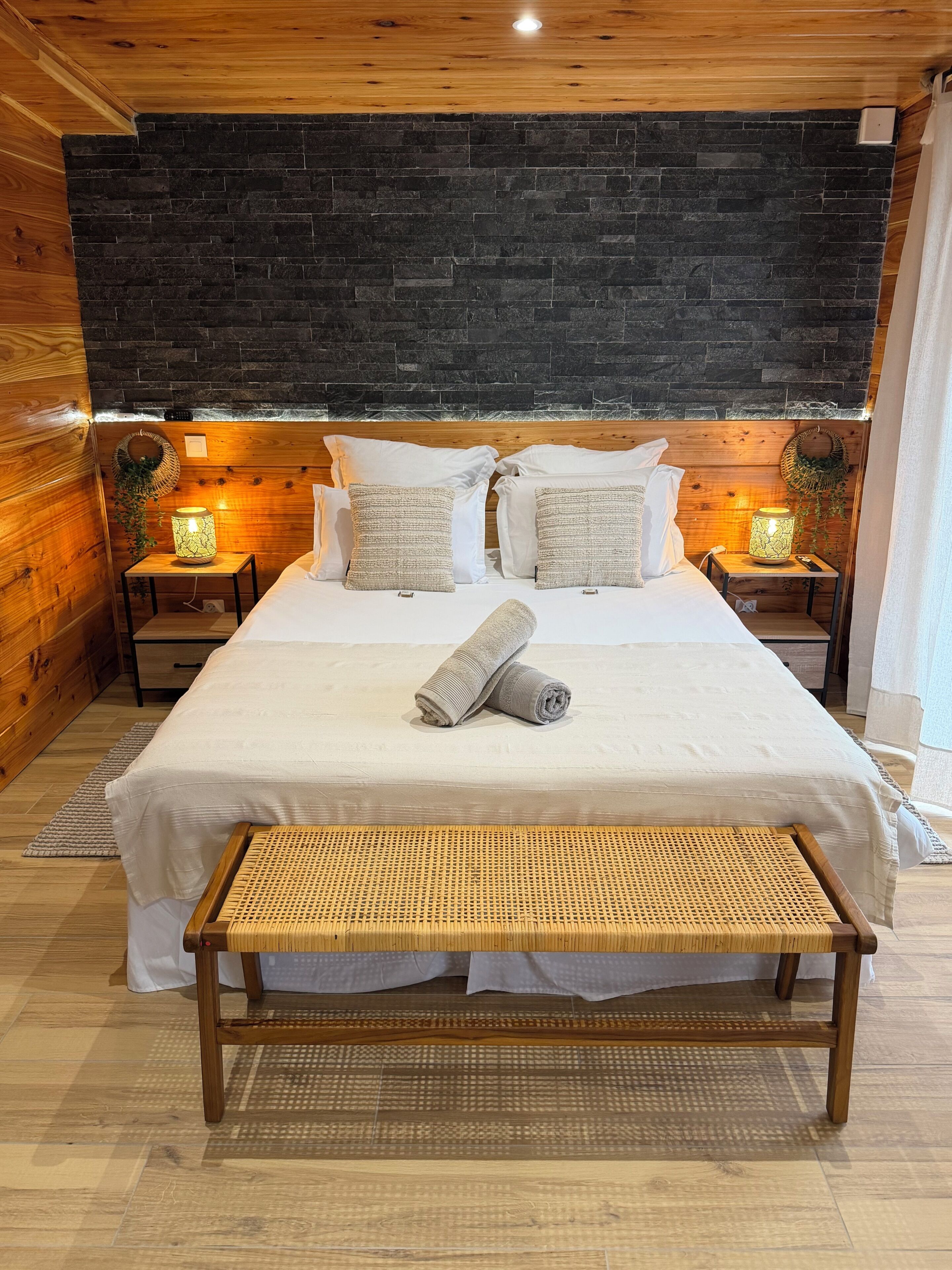 A bedroom with a large bed, two bedside tables, a bench, and a dark brick wall.