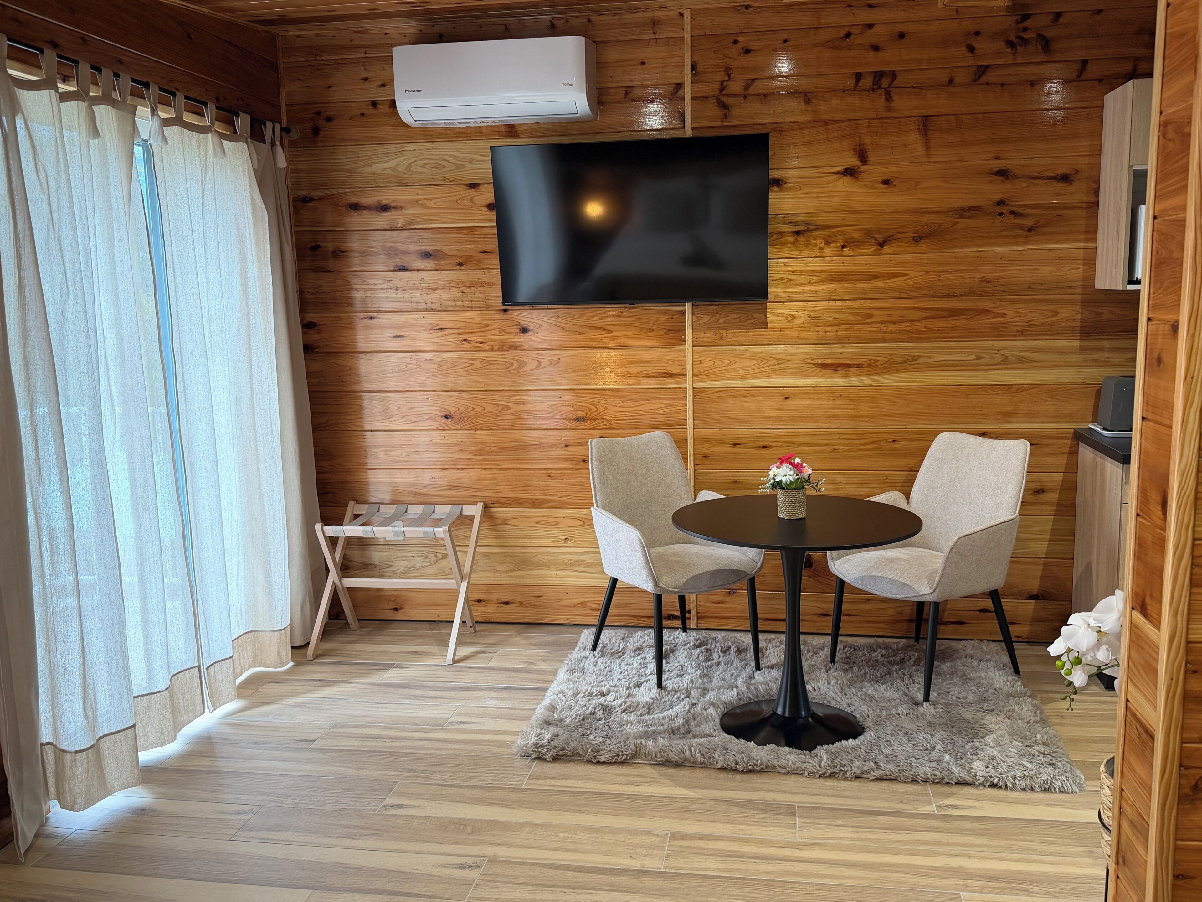 Una habitación con paredes de madera, un televisor de pantalla plana montado en la pared, dos sillas alrededor de una pequeña mesa redonda con un jarrón de flores, un taburete de madera y una alfombra mullida en el suelo.