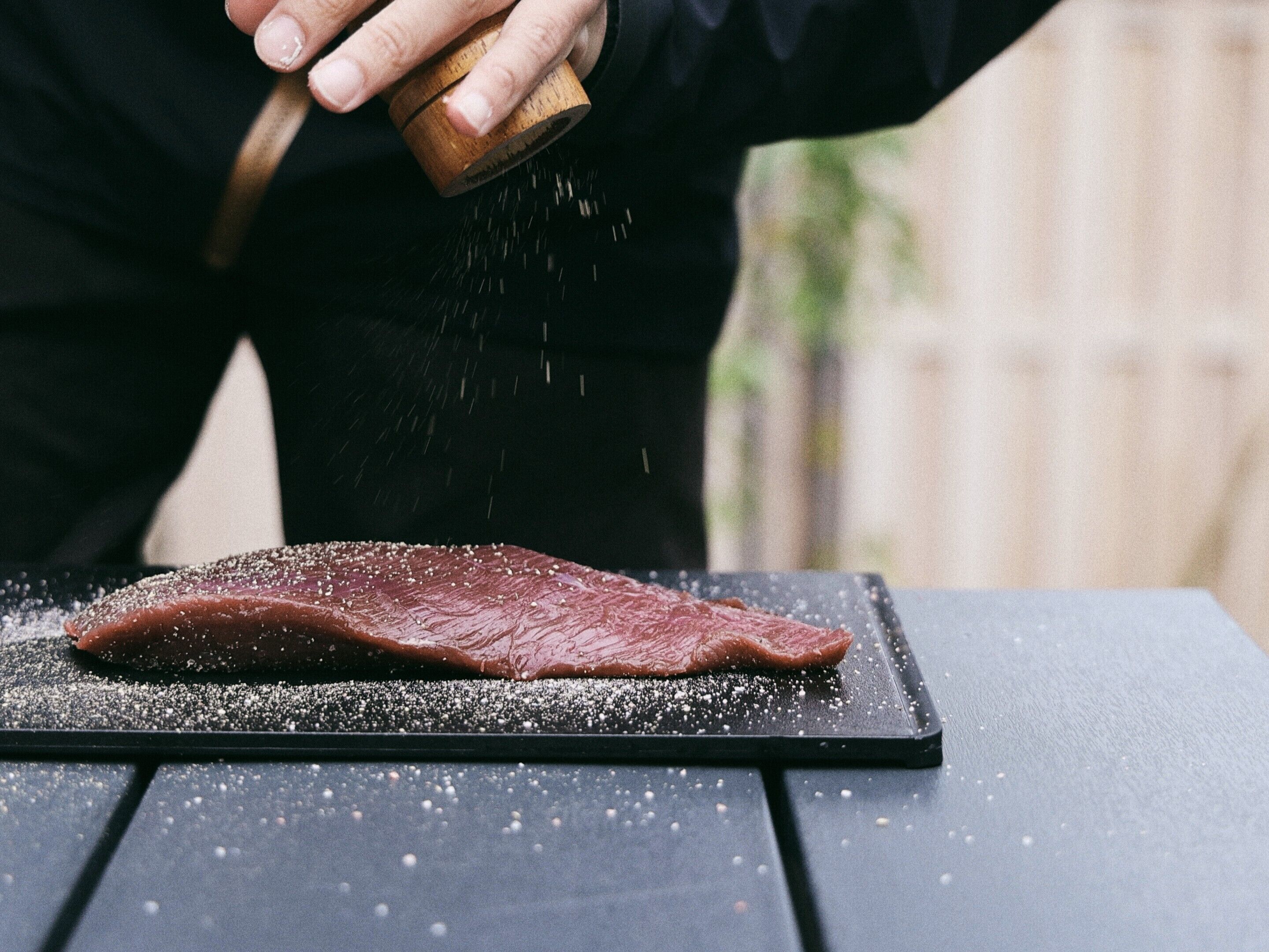 Iemand brengt kruiden aan op een steak op een zwart snijplank.