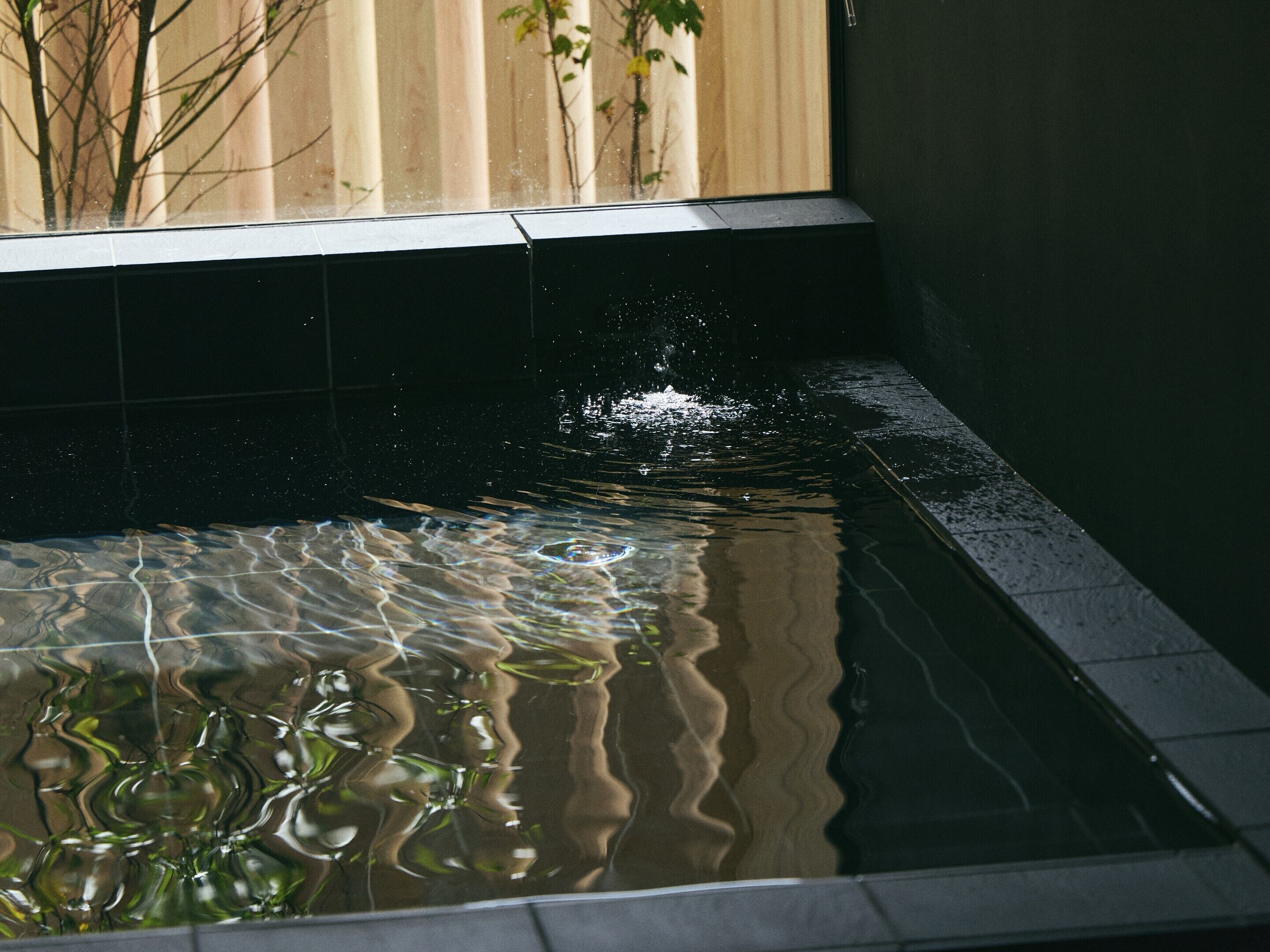 A clear pool with ripples and reflections, surrounded by a dark tiled edge and a wooden fence in the background.