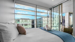 Ein modernes Schlafzimmer mit einem großen Bett, Blick auf die Stadt durch raumhohe Fenster und einem Balkon mit Ausblick auf die Häuser.