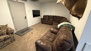 Ein Wohnzimmer mit einem braunen Ledersofa, einem aufgehängten Hirschkopf, einem weiteren braunen Ledersessel, einem gemusterten Sessel und einem Flachbildfernseher.