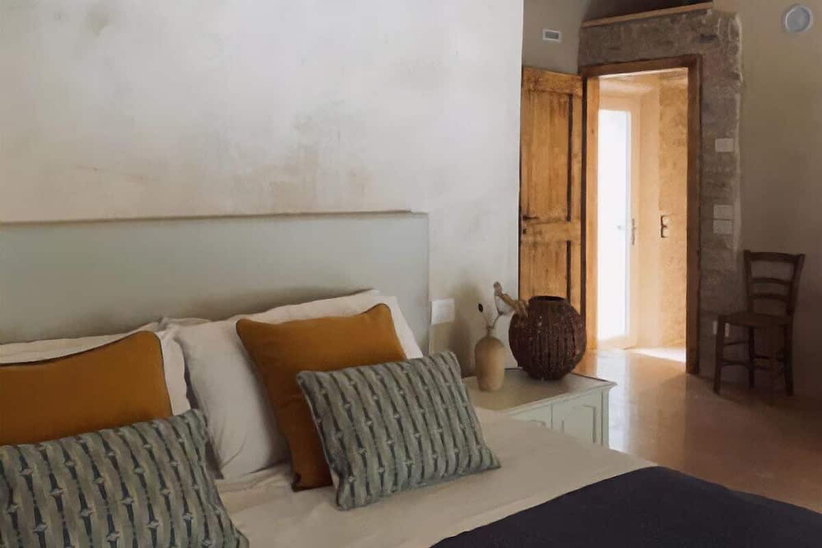 Um quarto com cama, almofadas, mesa de cabeceira, um vaso e uma cadeira.