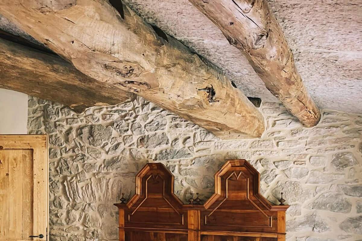 Quarto com cama de madeira, parede de pedra e vigas de madeira aparentes.