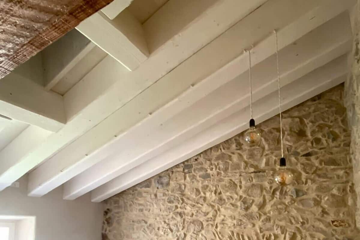 Quarto com parede de pedra, cama com roupa de cama azul e branca, mesa de cabeceira e luminária suspensa.