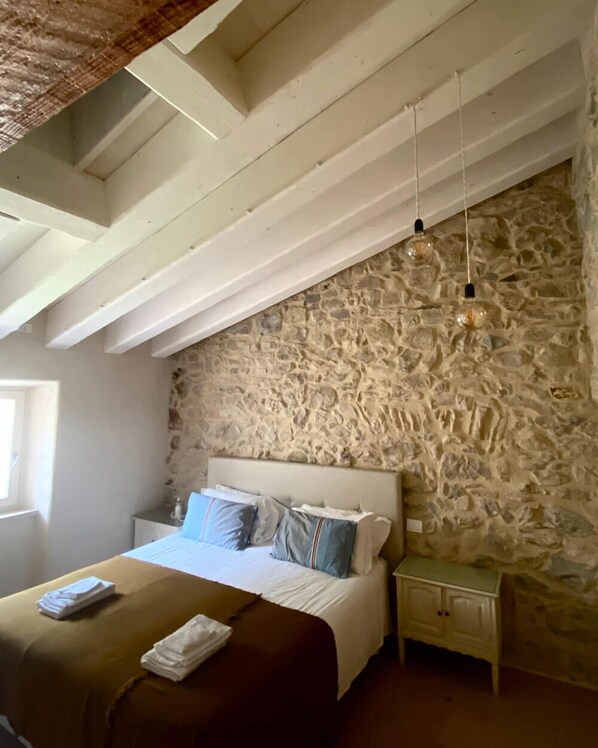 Un dormitorio con pared de piedra, una cama con ropa de cama azul y blanca, una mesita de noche y una lámpara colgante.