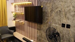 A modern room with a flat-screen TV mounted on a wooden panel wall, a small table with a bench, a potted plant, and a standing fan. - 🏡 Luxury 2 bedroom apartment (Lagos)