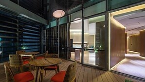 A modern interior with a round wooden table and wicker chairs, large glass doors leading to a bright room, and a visible outdoor area.