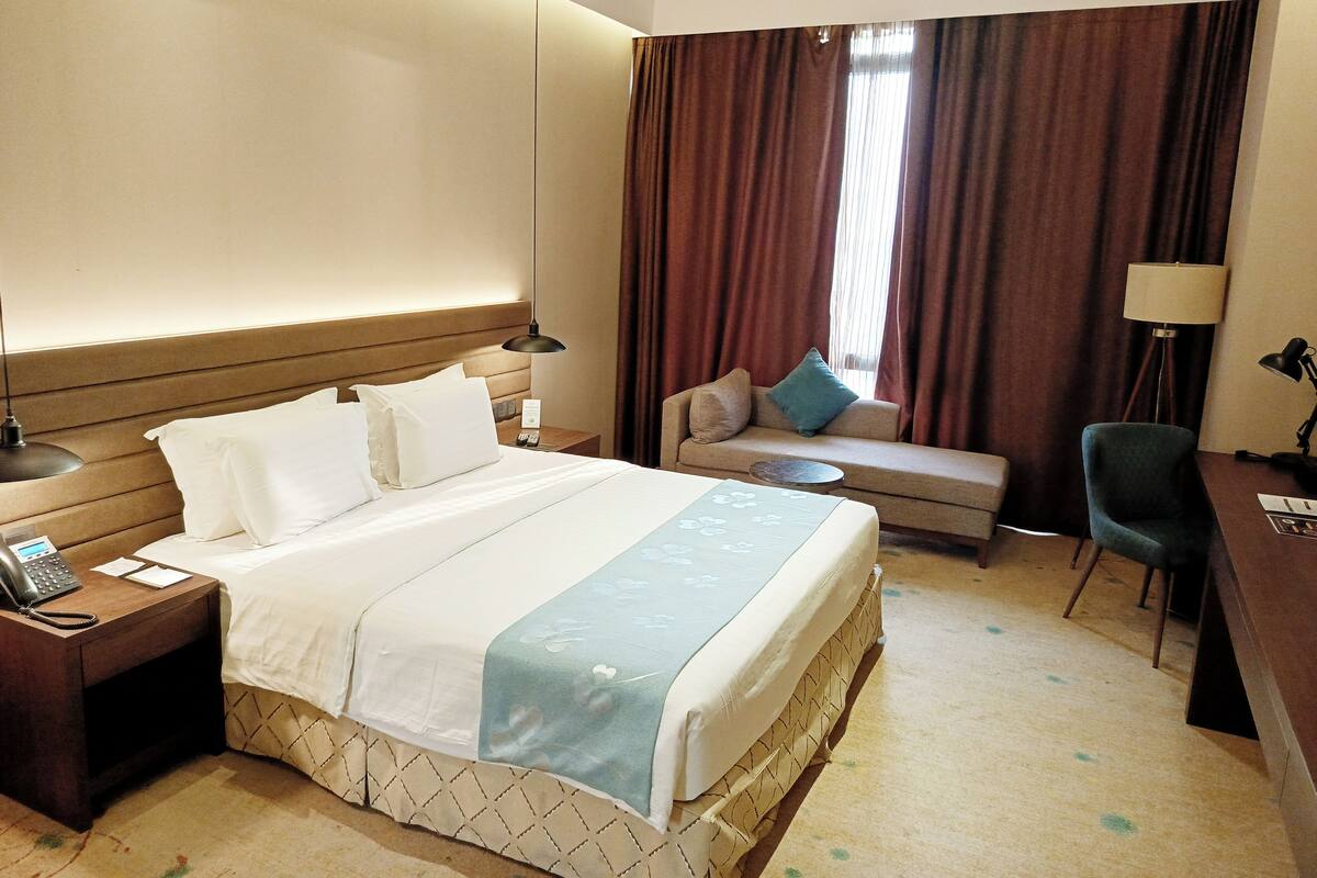 A hotel room with a bed, bedside table, lamp, and a sofa with a cushion.