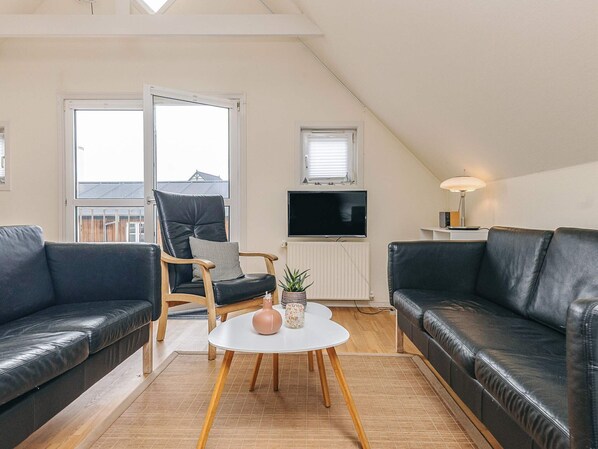 A modern living room with two black leather sofas, a wooden chair, a white coffee table, and a television mounted on the wall.