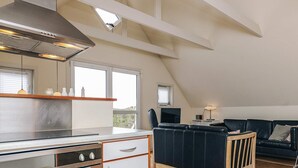 Een moderne keuken met een inbouwfornuis, een kleine koelkast en een houten aanrecht. In de woonkamer staat een zwarte leren bank en een houten stoel.