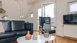 Een moderne woonkamer met een zwarte leren bank, een houten stoel met kussen, een klein wit tafeltje met decoratieve objecten en een groot raam met zonwering.