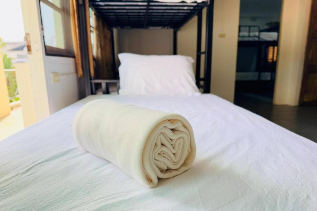 Una cama bien tendida con una toalla blanca enrollada en una habitación con una cama litera al fondo.