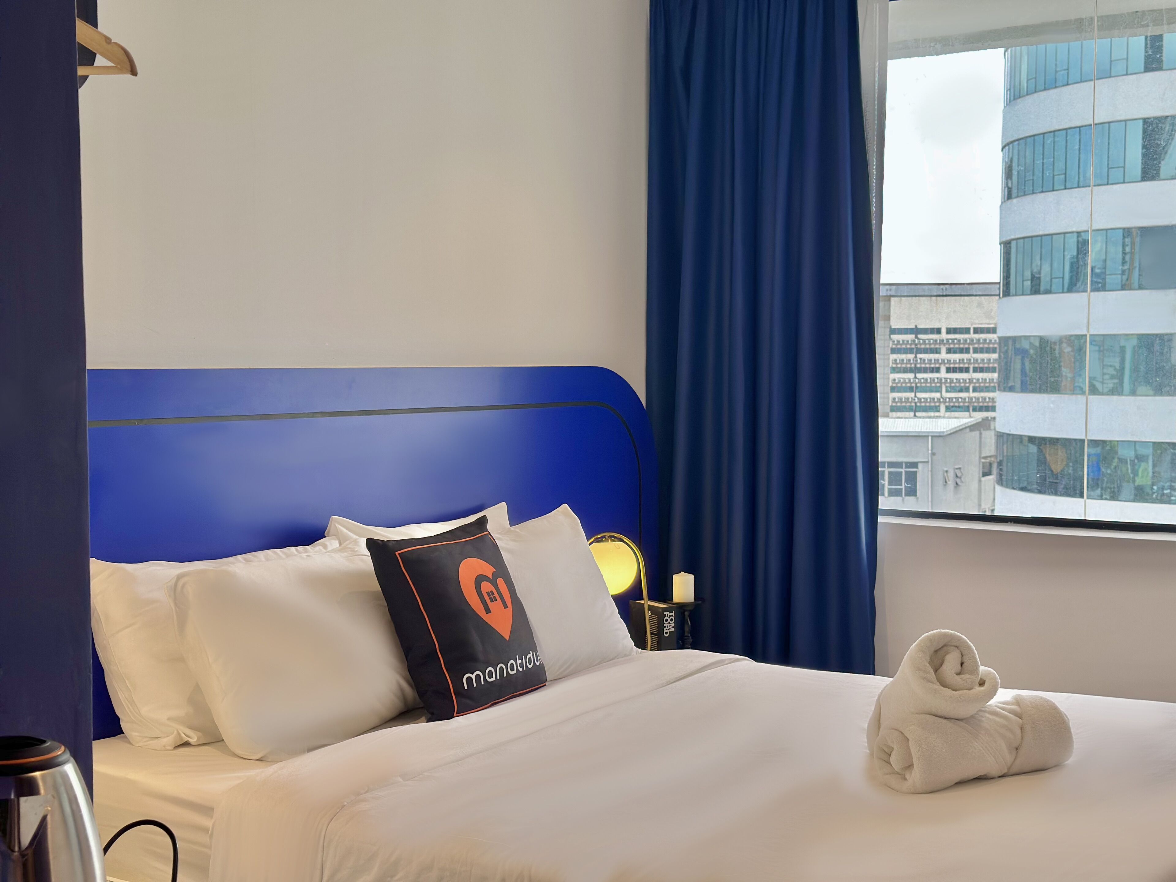 A hotel room with a blue headboard, white bedding, a pillow with a logo, a rolled towel, and a window with a city view.