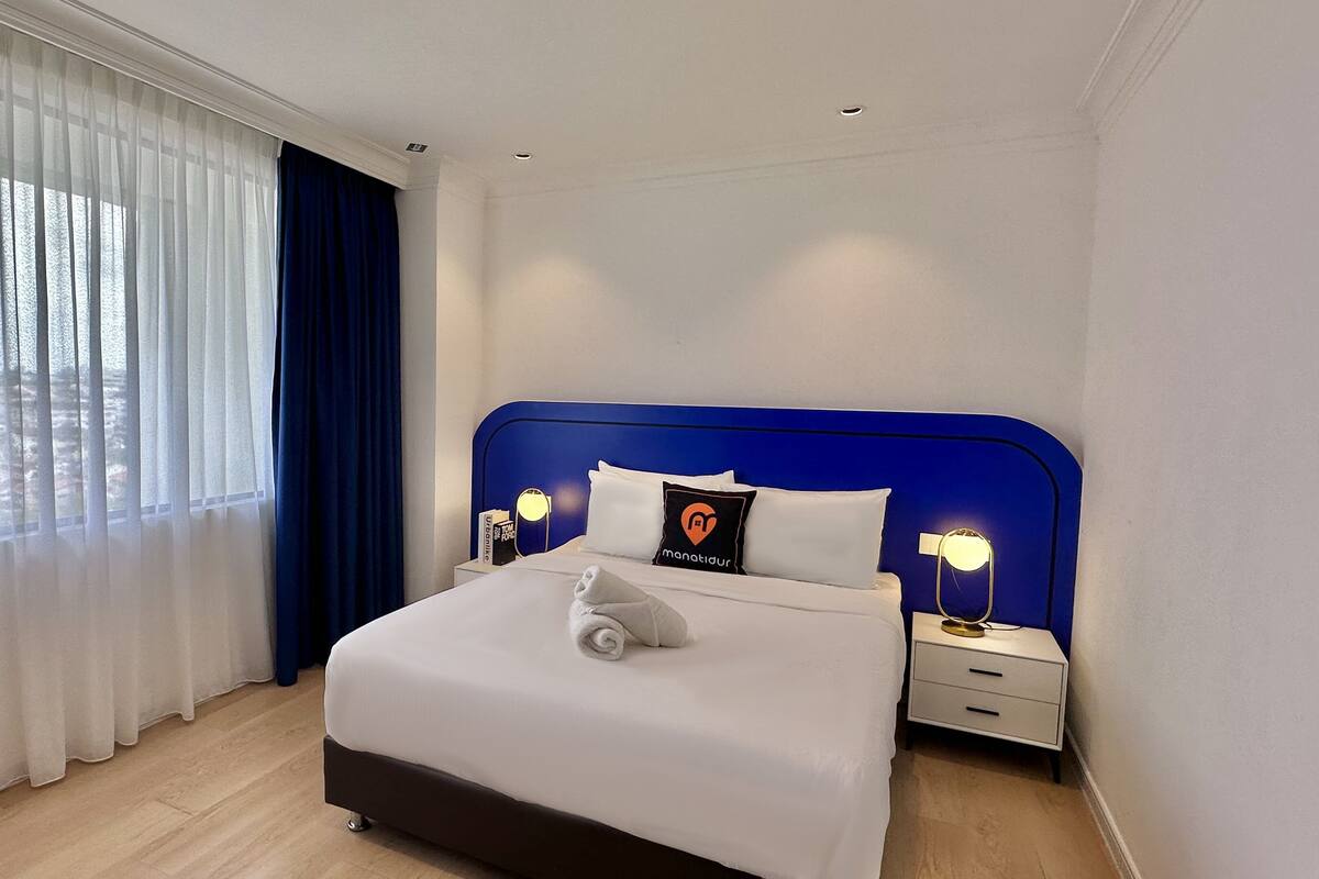 Ein Hotelzimmer mit Bett, Nachttisch, Lampe und einem fenster mit Vorhängen.