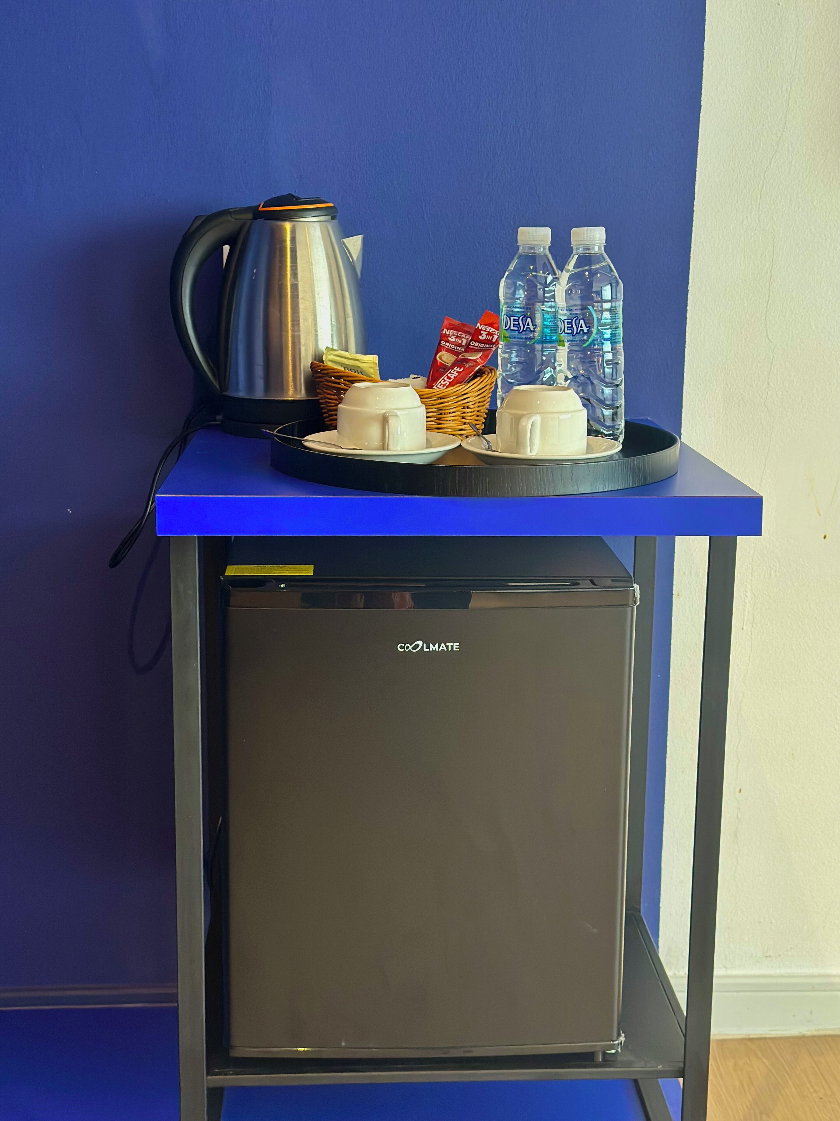 A small kitchenette with a coffee maker, two cups, a basket with a packet, and two water bottles on a blue tray.