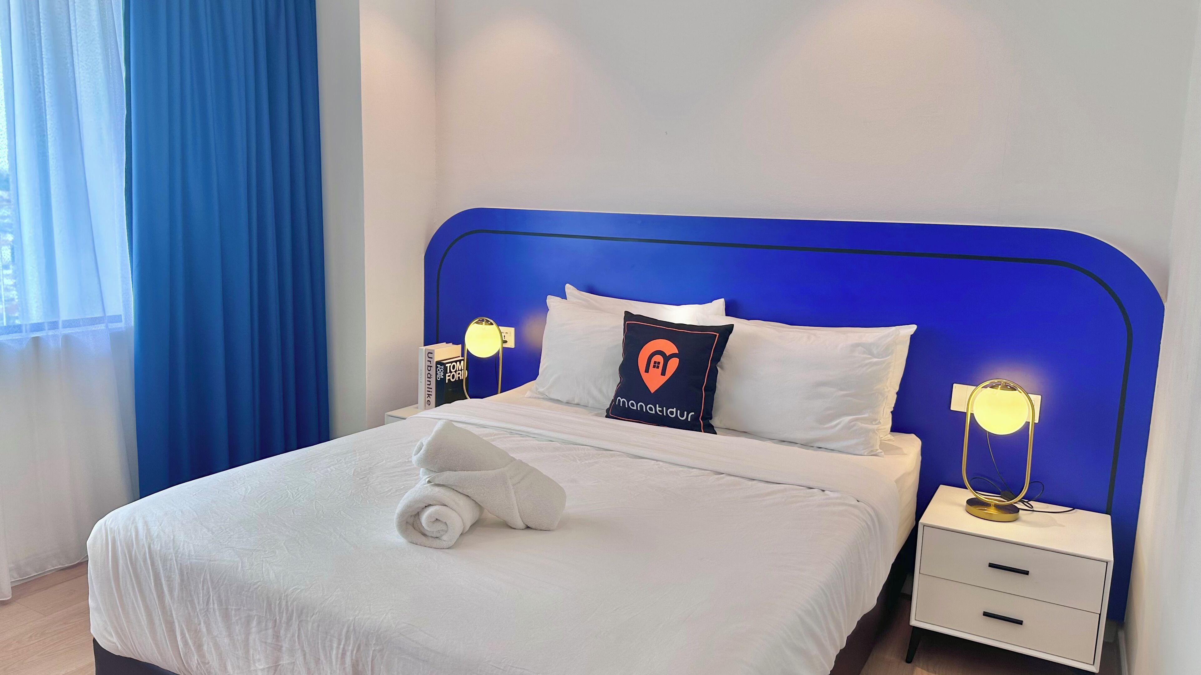 Ein ordentlich gemachtes Bett mit weißen Bettwäsche und einem zusammengerollten Handtuch, ein blauer Kopfteil, zwei Nachttische mit Lampen und ein Fenster mit blauen Vorhängen.