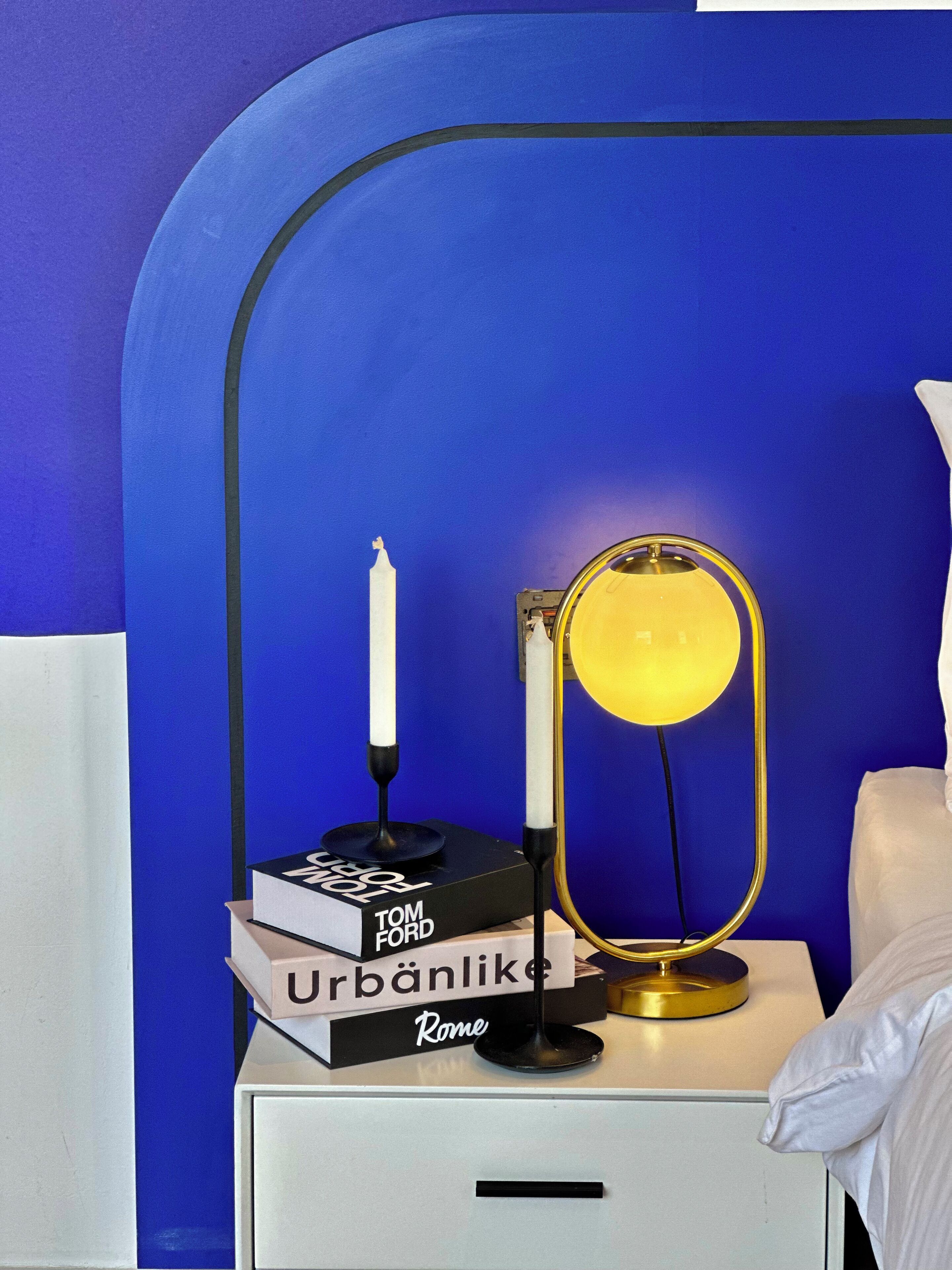 A neatly arranged bedside table with a lamp, a book titled 'Urban Like' by Tom Ford, and a candle.