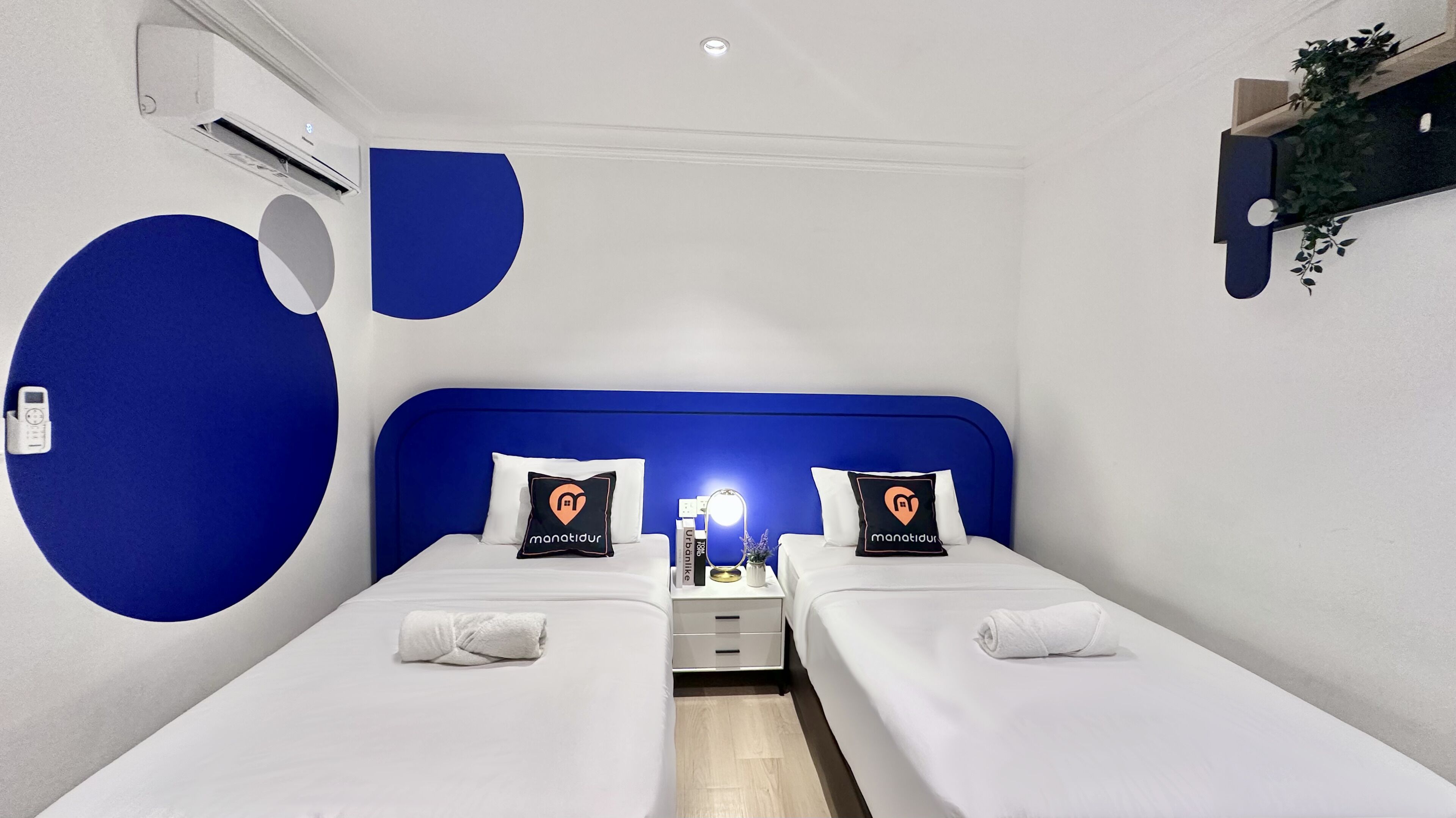 A hotel room with two single beds, a bedside table with a lamp, and a wall-mounted shelf with a plant.