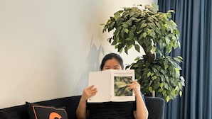 Uma pessoa sentada num sofá com um livro, uma planta e uma vela sobre uma mesa.