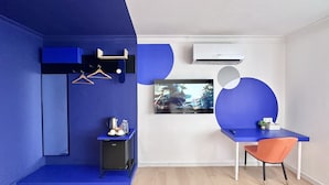 Quarto moderno com parede azul de destaque, uma pequena escrivaninha com cadeira, TV na parede e uma cozinha compacta.