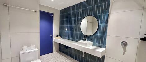 Un baño moderno con inodoro blanco, puerta azul, lavamanos blanco con espejo encima y cabezal de ducha.
