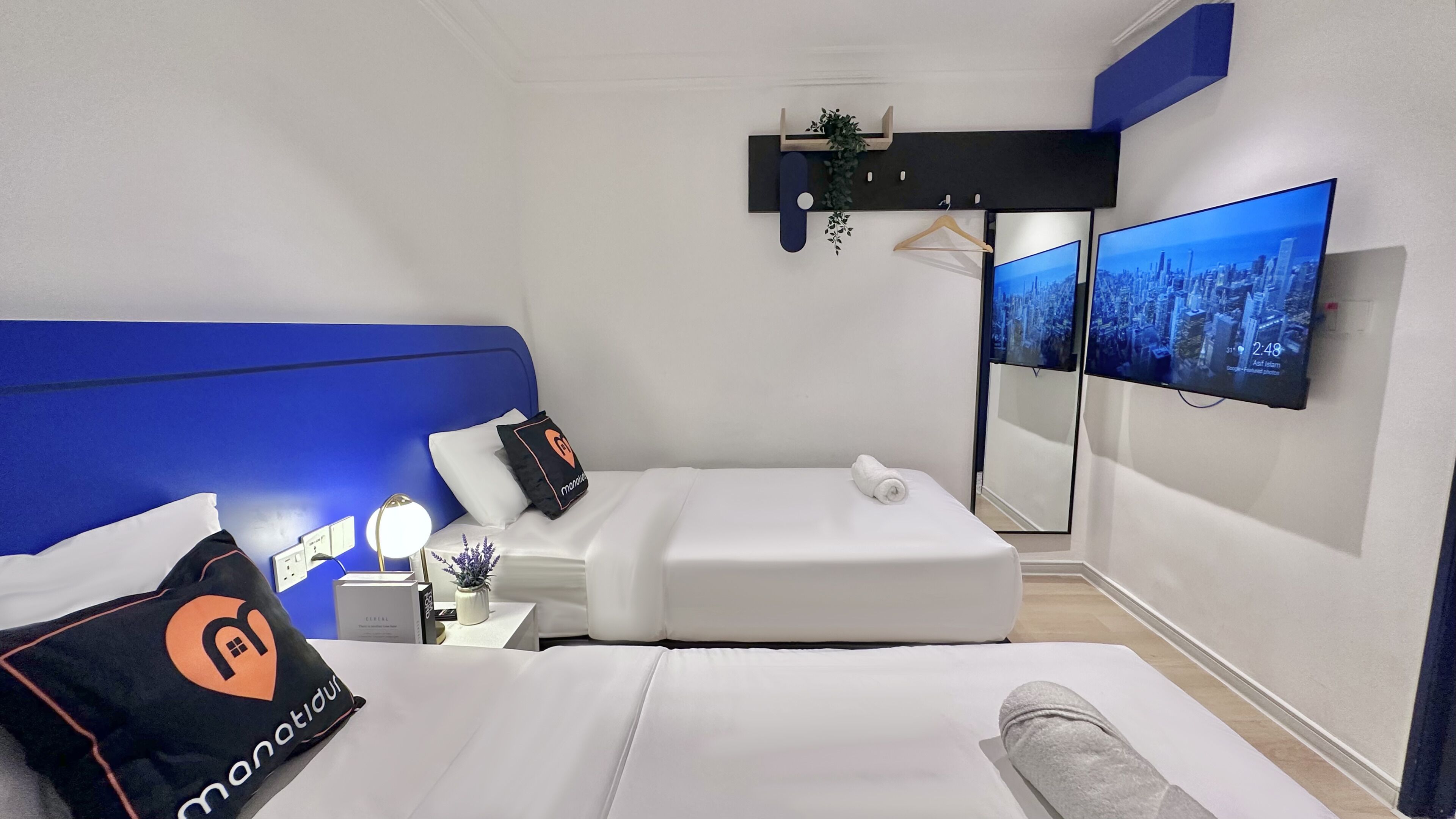 Ein modernes Hotelzimmer mit einem großen Bett, einem Nachttisch mit Lampe, einem kleinen Beistelltisch, einem Spiegel und einem an der Wand befestigten Fernseher, der eine Stadtansicht zeigt.