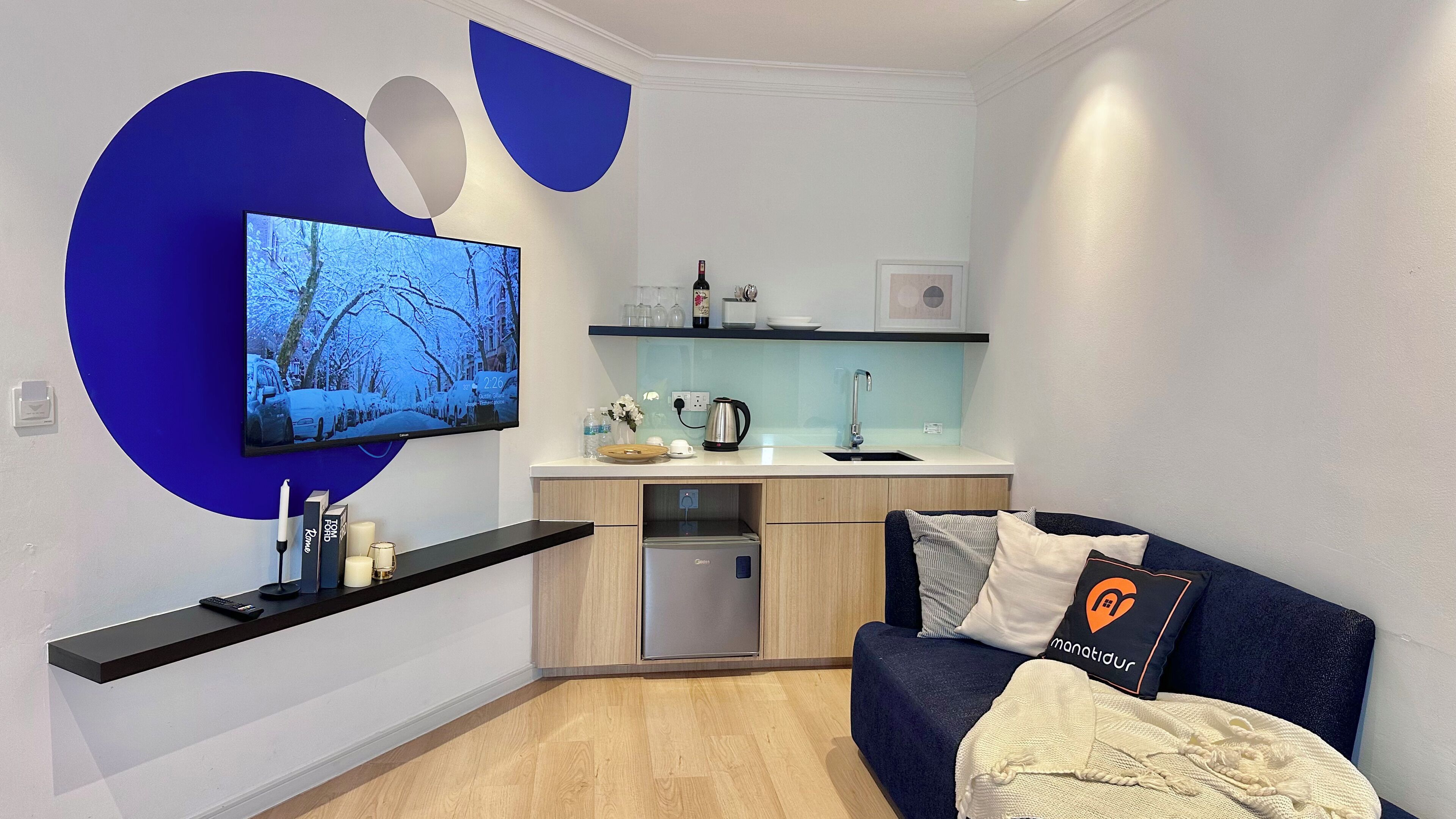 A modern living space with a kitchen area, a flat-screen TV mounted on the wall, and a sofa with decorative pillows.