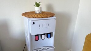 Dispensador de agua blanca con una planta pequeña en la parte superior, situado en un rincón de una habitación con suelo estampado.