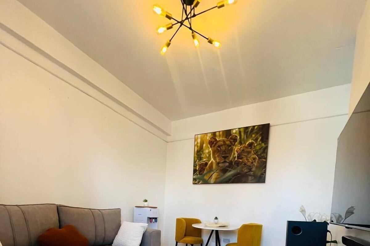 A modern living room with a grey sofa, yellow throw blanket, and a painting of lions. There is a small table with chairs and a white cabinet.